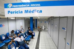 Sala de espera de uma agência da Previdência Social com placa indicativa de Perícia Médica no teto e pessoas sentadas aguardando atendimento para verificar acidente
