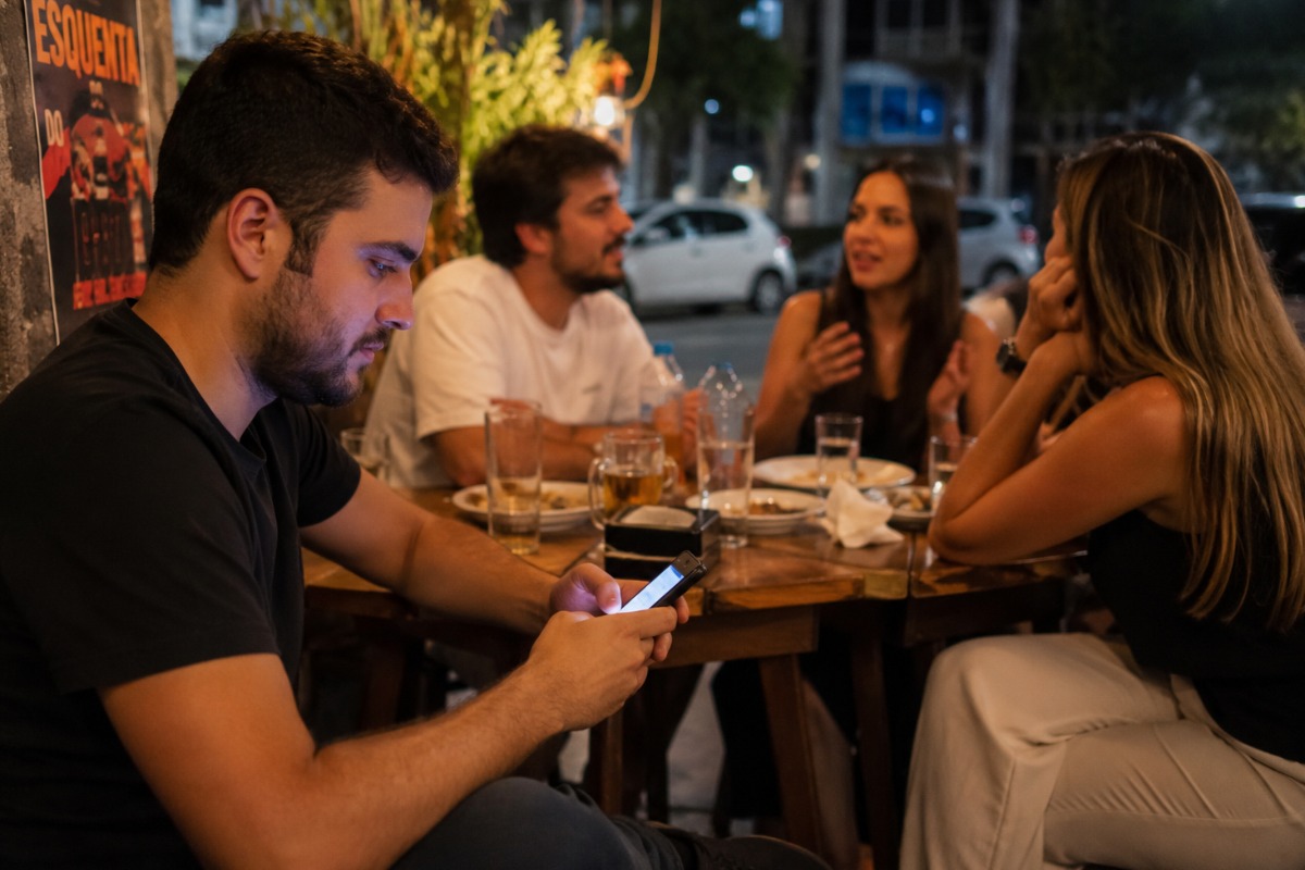 Pessoa olha para o celular enquanto amigos conversam ao redor de uma mesa