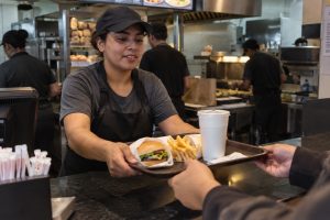 Atendente entrega bandeja com lanche em balcão de restaurante de fast food, com cozinha e funcionários ao fundo
