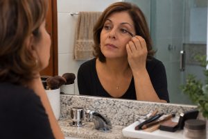 Mulher com mais de 50 anos aplica lápis próximo aos cílios diante do espelho, com outros tons de lápis sobre a bancada