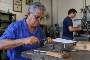 Profissional com cerca de 60 anos realiza tarefa cuidadosa no trabalho enquanto colega mais jovem aparece ao fundo