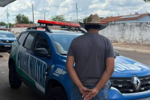 Homem de costas, usando chapéu, camiseta cinza e calça jeans, em frente a uma viatura da Polícia Militar estacionada em área urbana