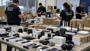 Produtos eletrônicos apreendidos aparecem sobre mesas enquanto agentes realizam fiscalização das mercadorias