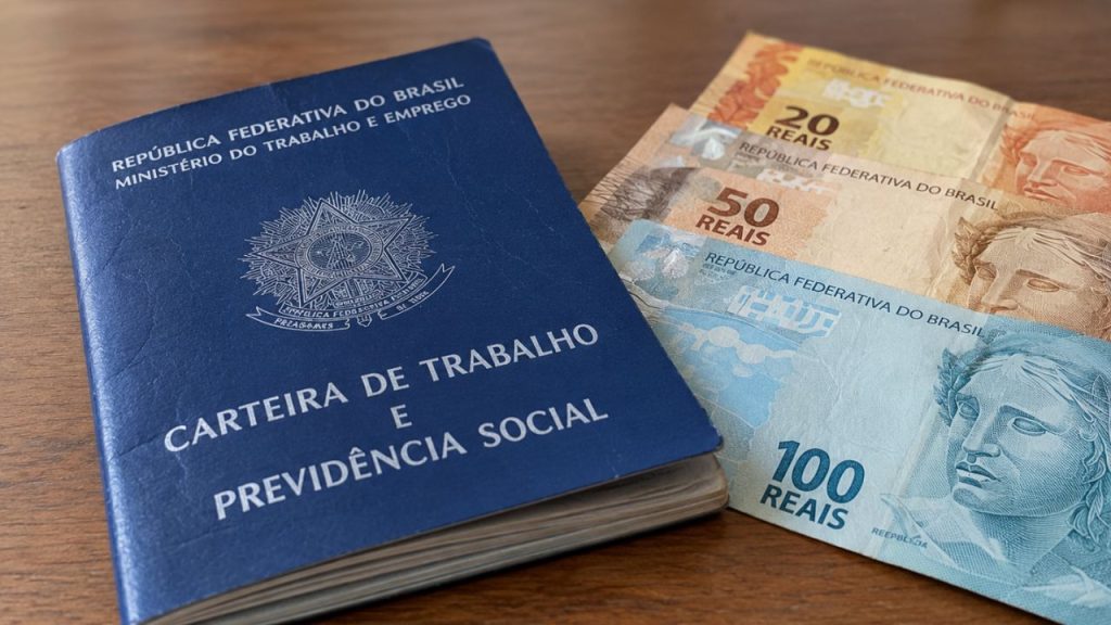 Carteira de Trabalho aparece ao lado de cédulas de real sobre uma mesa, representando pagamento de benefício trabalhista