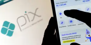 Mão tocando a tela de um aplicativo bancário em um celular, com opções de Pix, empréstimo e cartão, diante do logotipo do Pix desfocado ao fundo.